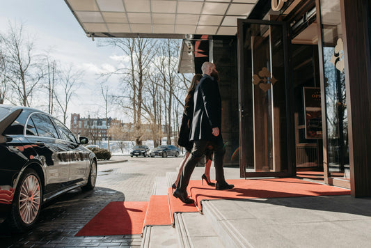 Two well-dressed people walking up steps to enter a hotel with a luxury car parked nearby