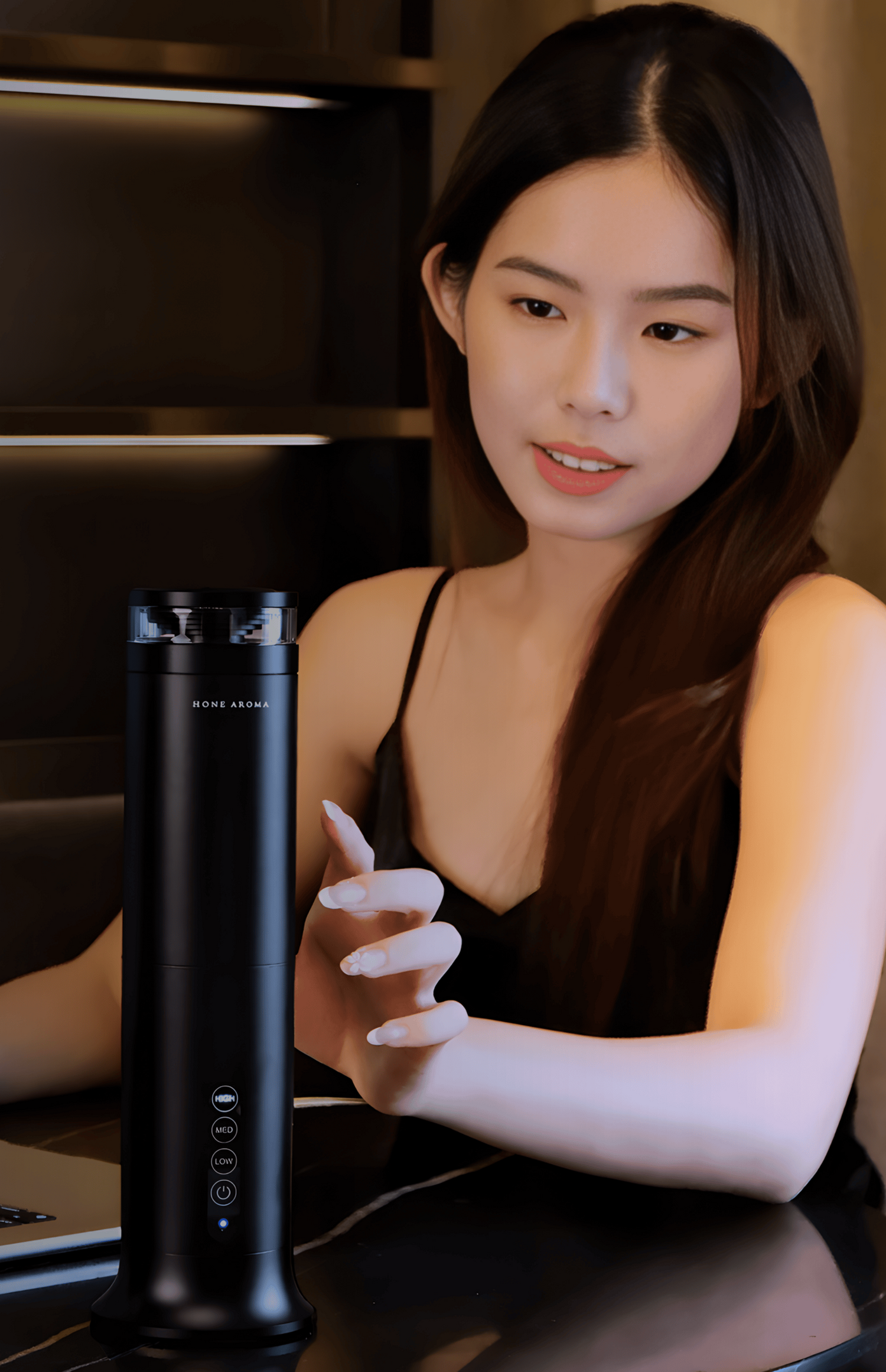 Woman sitting at a table with a laptop and a Pillar Scent Diffuser in a dimly lit room.